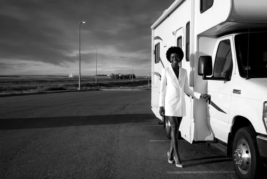 Black-and-white generated image: the guide's character stands by a white motorhome on a wide paved lot at dusk, the prairie and a highway behind her.
